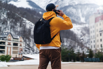 Back view. Man tourist in yellow hoodie with black backpack stands on background of high snowy mountains, talking on mobile phone.