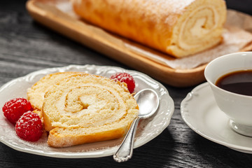 Ciasto - rolada biszkoptowa z kremem mascarpone na talerzyku, obok kawa w filiżance, wszystko postawione na czarnym drewnianym blacie © Grzej