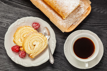 Ciasto - rolada biszkoptowa z kremem mascarpone na talerzyku, obok kawa w filiżance, wszystko postawione na czarnym drewnianym blacie