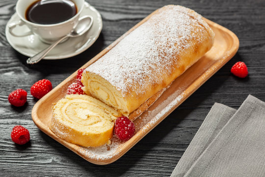 Ciasto - rolada biszkoptowa z kremem mascarpone na drewnianej tacce, obok kawa w filiżance, wszystko postawione na czarnym drewnianym blacie