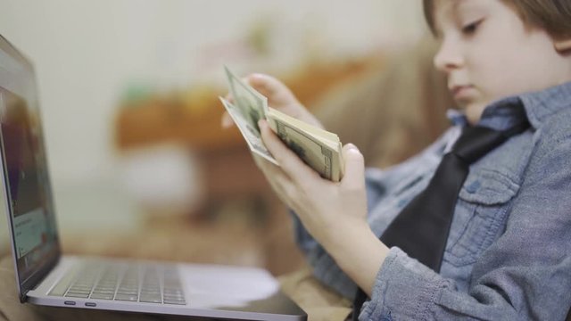 The little boy in jeans jacket sitting on sofa in front of laptop counting the money. A bundle of dollars in child's hands. The leisure of modern kid. Cute son of rich parents