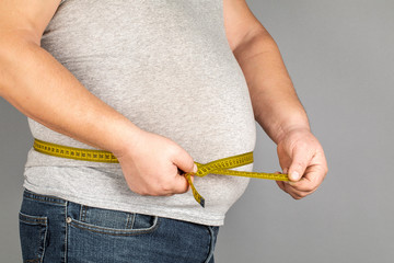 A man measures his fat belly with a measuring tape. on a gray background.