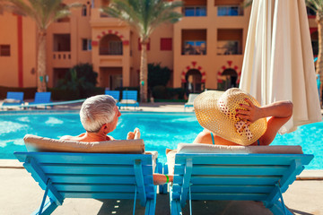 Senior couple relaxing by swimming pool lying on chaise-longues. People enjoying summer vacation. - obrazy, fototapety, plakaty
