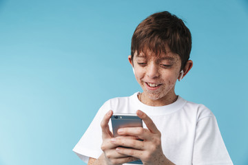 Image of joyful caucasian boy 10-12y with freckles wearing white casual t-shirt smiling and holding smartphone