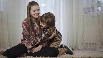 Young girl sit on the fluffy rug in the living room and her brother come to cuddle her. Family love and happiness concept.