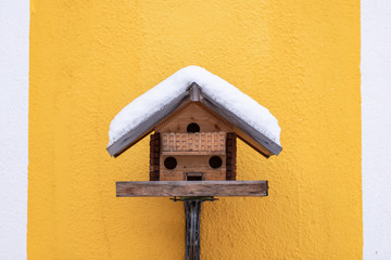 Homemade wooden bird's feeder in winter, under the snow on the yellow wall background.