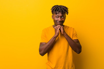 Young black man wearing rastas over yellow background keeps hands under chin, is looking happily...