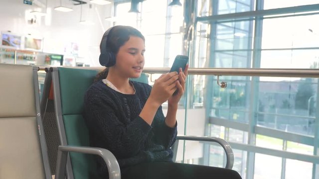 airport waiting hall room for a flight by plane. young happy teenage girl in headphones listening to the music on smartphone chatting communicates in the messenger. teenager girl in social networks