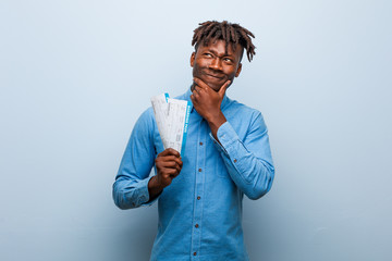 Young rasta black man holding an air tickets looking sideways with doubtful and skeptical expression.