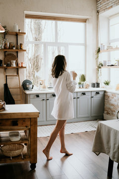Successful and confident woman on a morning coffee break, resting and enjoying the drink. Young girl in white panties and long shirt with naked shoulder and chest stand on the kitchen of the house
