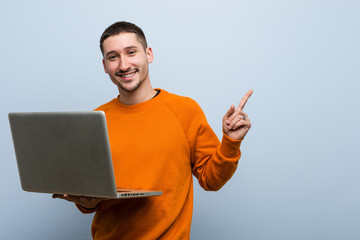 Young caucasian man holding a laptop smiling cheerfully pointing with forefinger away.