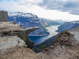 A man wearing blue jacket sits at the hanging rock formation, Trolltunga with a view on Ringedalsvatnet lake, Norway. Slopes of the mountains are partially covered with snow. Freedom and happiness