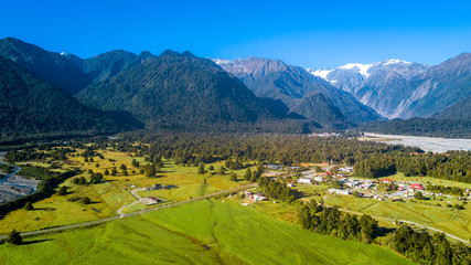 Naklejka na ściany i meble Small village in the middle of sunny valley with snowy mountains on the background. West Coast, South Island, New Zealand