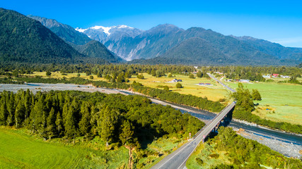 Fototapeta premium Bridge across the river in the middle of sunny valley with snowy mountains on the background. West Coast, South Island, New Zealand