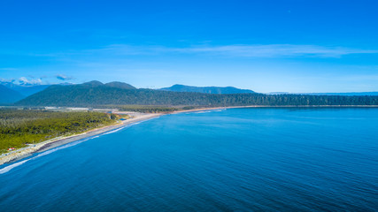 Road running along a sunny beach with a peninsula and mountains on the background. West Coast, South Island, New Zealand