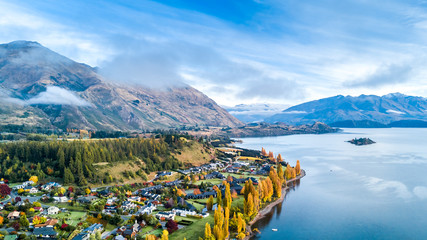 Small town surrounded by yellow autumn trees on a shore of pristine lake with mountains on the background. Wanaka, Otago, South Island, New Zealand - obrazy, fototapety, plakaty