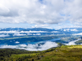 A view on the lake from a hill. In the back endless chains of Alps. Millstatter See in the valley bottom. Lots of clouds in the valley. Green slopes of the mountains.