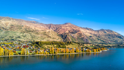 A small town surrounded by yellow autumn trees on a shore of a pristine lake with mountains in the...