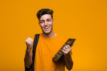Young student man holding a calculator celebrating a victory or success - obrazy, fototapety, plakaty