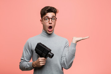 Young intellectual man holding a film camera impressed holding copy space on palm.