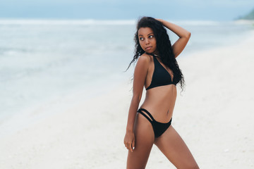 Fototapeta na wymiar Beautiful sexy dark skinned girl in swimwear posing on white sand beach, Afro american woman resting on paradise island
