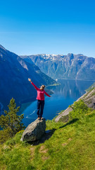 A girl in pink jacket stands on a rock with hands up in the air. Majestic view on Eidfjord from Kjeasen, Norway. Slopes are overgrown with green grass. Water has dark blue color. Freedom and happiness