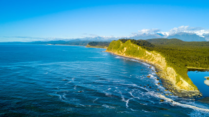 Fototapeta premium Remote rocky coastline with native forest and snowy mountain peaks on the background. West Coast, South Island, New Zealand