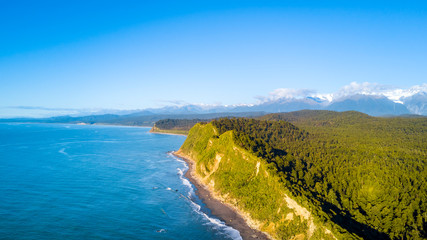 Remote rocky coastline with native forest and snowy mountain peaks on the background. West Coast, South Island, New Zealand