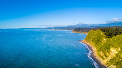 Fototapeta na wymiar Remote rocky coastline with native forest and snowy mountain peaks on the background. West Coast, South Island, New Zealand