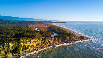 Fototapeta premium Sunset over remote beach on the coast on Tasman sea with native forest and mountains on the background. West Coast, South Island, New Zealand