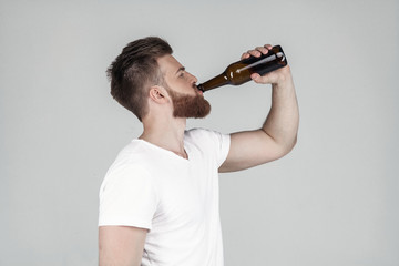 Portrait of a beautiful sexy bearded man dressed in a white T-shirt standing in profile and drinking beer, standing in front of a white background