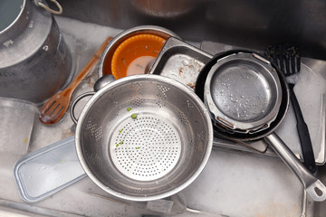 Closeup stack of dirty utensils in metal square sink at professional restaurant kitchen: stack of pans, colander, shovels faucet, dishes detergent, sponge. Top view