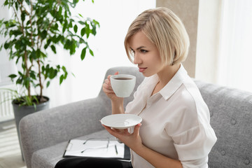 Young beautiful blonde girl with short hair in a white shirt and glasses is sitting on the sofa in bright in the office against the window. Drinking coffee or tea in a working break.