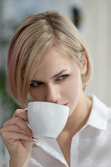 Young beautiful blonde girl with short hair in a white shirt and glasses is sitting on the sofa in bright in the office against the window. Drinking coffee or tea in a working break.