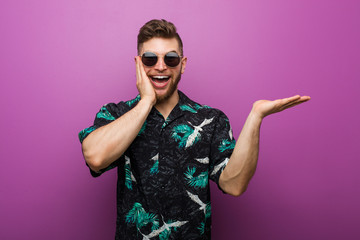 Young man wearing a vacation look holds copy space on a palm, keep hand over cheek. Amazed and delighted.