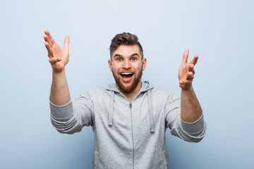 Young handsome fitness man receiving a pleasant surprise, excited and raising hands.