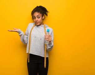 Young fitness black woman doubting and shrugging shoulders.Holding a water bottle.