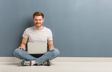 Young redhead student man sitting on the floor cheerful with a big smile. He is holding a laptop.