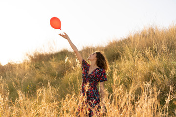 Happy young woman standing in summer meadow, letting go of a red balloon