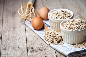 Fresh chicken eggs, oat flakes in ceramic bowls and wooden spoon on rustic wooden table background.