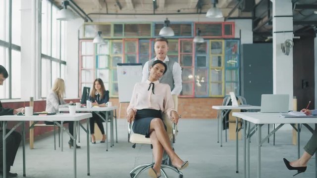 A young businessman is walking around the office, an African American woman employee rolls out to meet him on a chair, he picks it up and cheers her around the office. Office life. Creative interior
