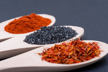 Saffron, Black Lava Sea Salt and Sweet Paprika in wooden spoons on dark background.