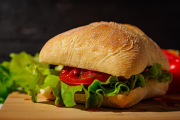 VEGAN SANDWICH WITH RED TOMATO GREEN SALAD LEAVES AND TOMATO SAUCE IN CIABATTA BREAD LAY ON THE CUTTING BOARD ON THE BLACK BACKGROUND FRONT VIEW