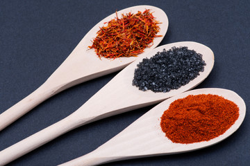 Saffron, Black Lava Sea Salt and Sweet Paprika in wooden spoons on dark background.