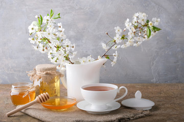 A cup of tea, a white kettle with cherry blossoms, jars and a saucer with honey on the table. Morning breakfast.