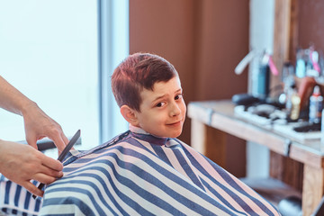 Cute little child is getting trendy haircut from barber at busy barbershop.