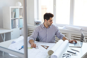 Portrait of young engineer working with plans and blueprints sitting at desk in office