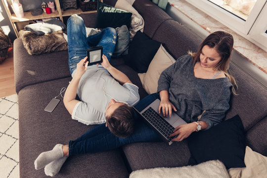 Young couple relaxing on sofa and using notebook, smartphone and ebook reader