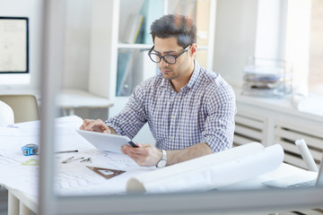 Portrait of engineer using digital tablet while sitting at desk in office behind glass, copy space
