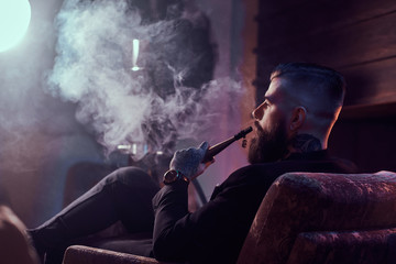 Obraz premium Attractive bearded man is making nice misty vapour while relaxing near fireplace and smoking hookah.